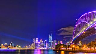 An image of the Sydney Harbour lit up with a purple and blue colour scheme for Vivid, including the Sydney Harbour Bridge and Sydney Opera House.