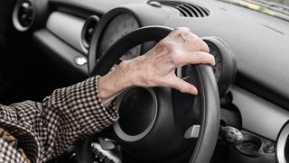An older person's hand resting on a steering wheel.