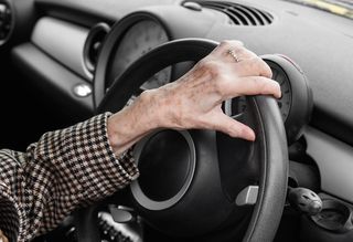 An older person's hand resting on a steering wheel.