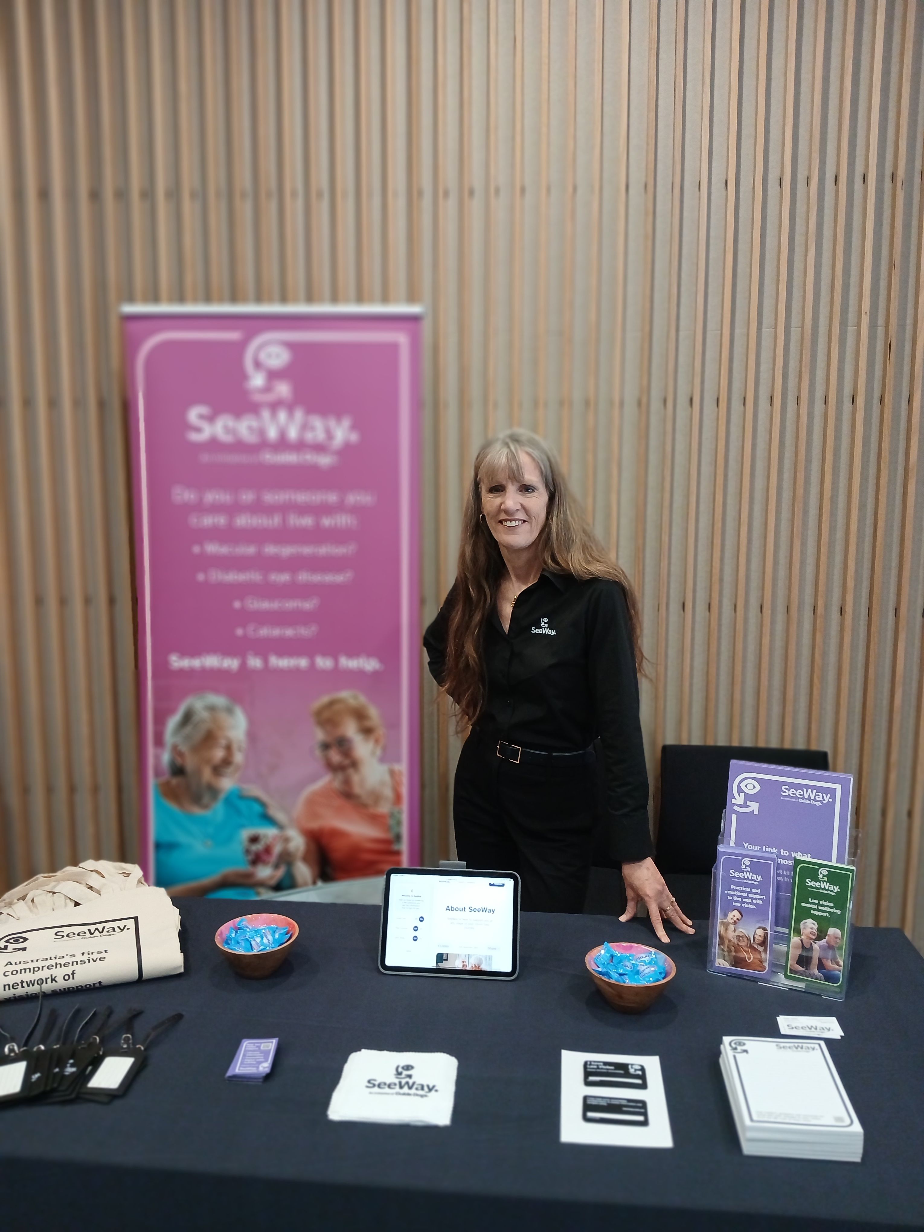Kaye Jones posing behind a table covered in SeeWay collateral, standing next to a pink SeeWay banner.