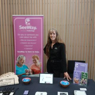 Kaye Jones posing behind a table covered in SeeWay collateral, standing next to a pink SeeWay banner.