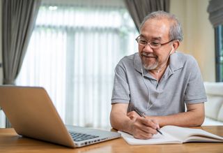 Older person using laptop with headphones while writing in a notebook