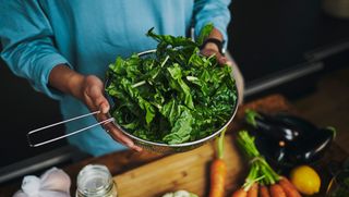 A person holding a strainer filled with assorted leafy green vegetables.