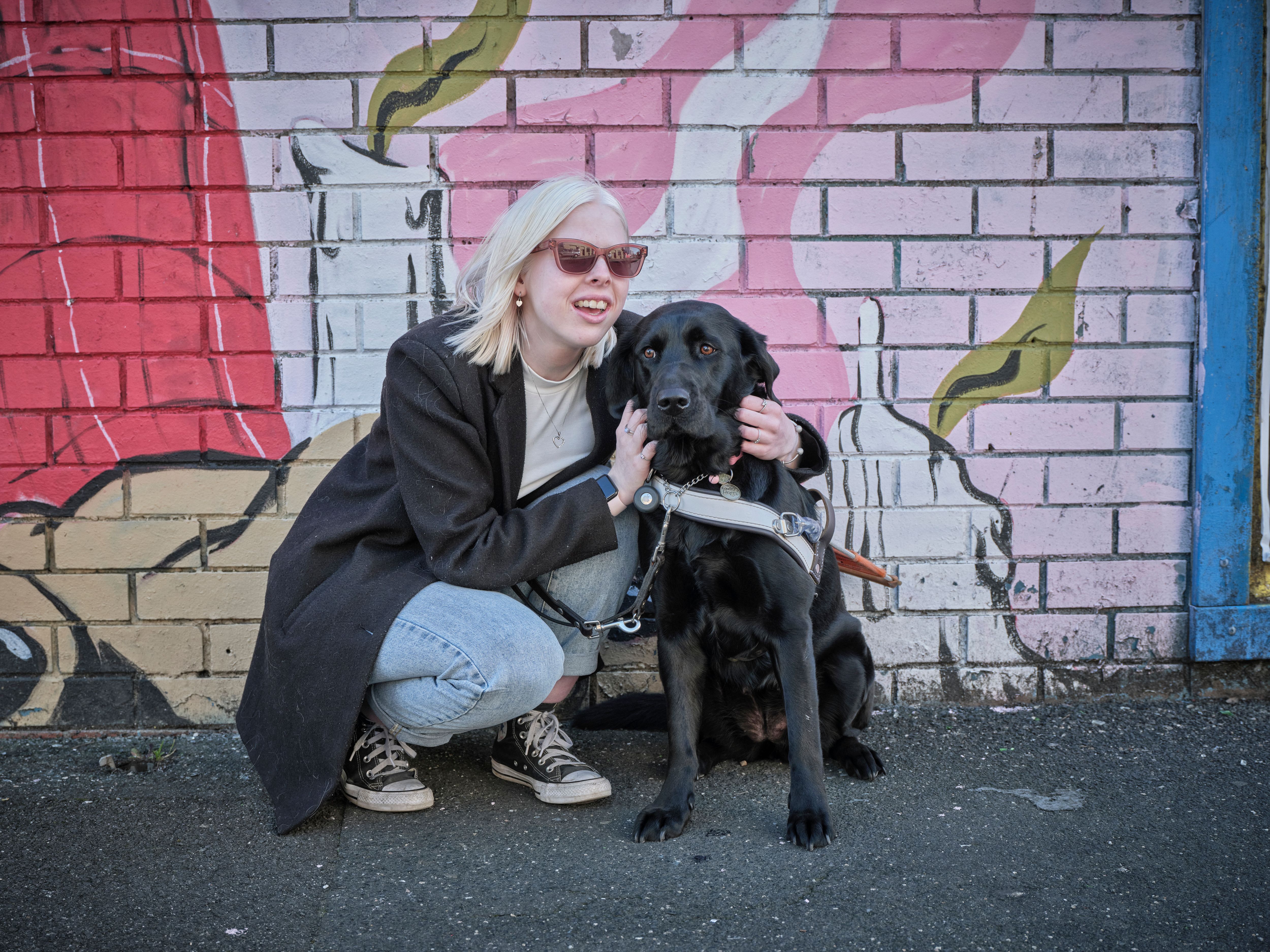 Lucy crouched in front of a brightly coloured mural with her black Guide Dog Dottie.