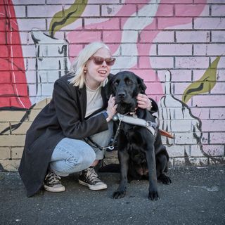 Lucy crouched in front of a brightly coloured mural with her black Guide Dog Dottie.
