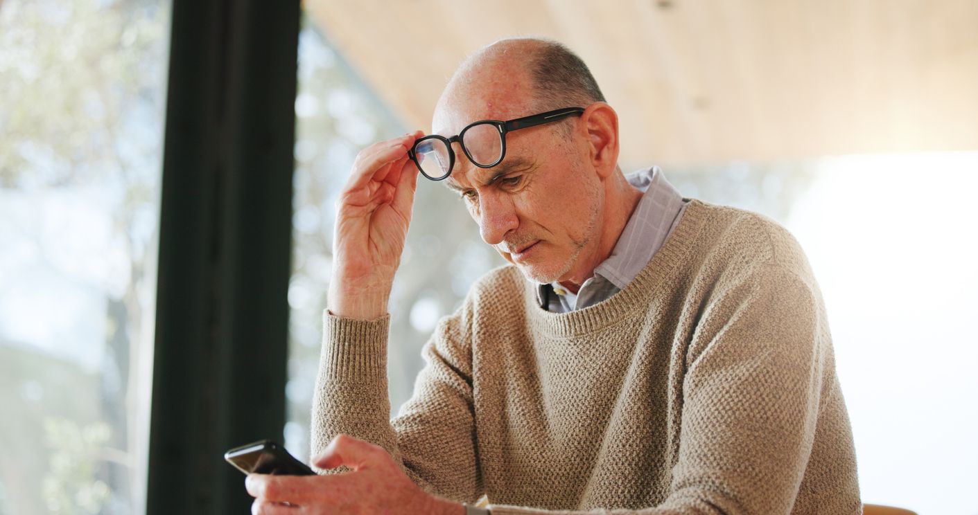 A man lifts his glasses to look at his phone screen