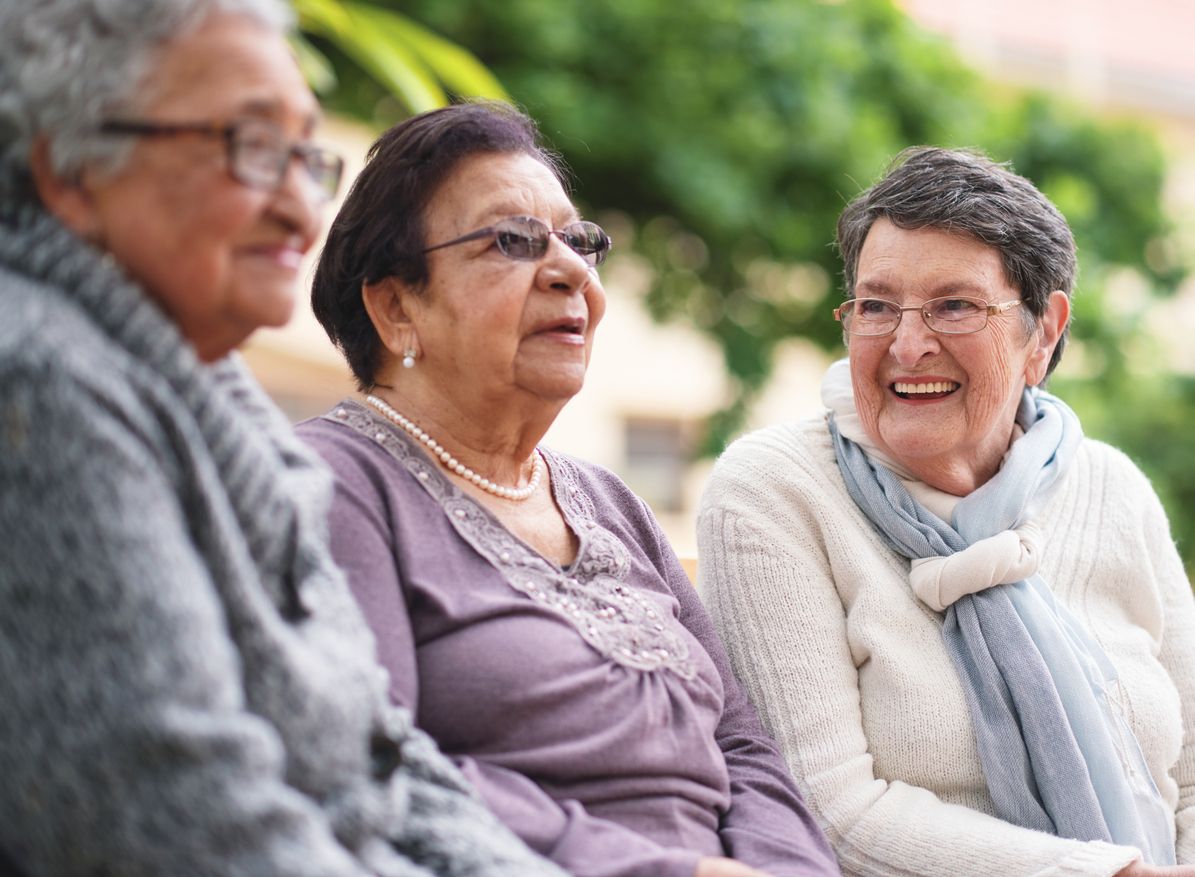 Three smiling older women sitting together on a bench outside.
