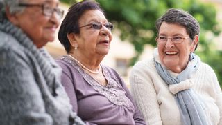 Three smiling older women sitting together on a bench outside.