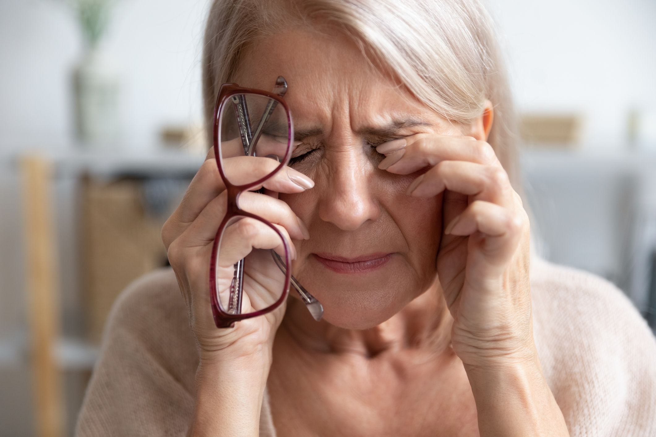 An older woman with her eyes closed in discomfort, rubbing them with her fingers while holding a pair of glasses in one hand.