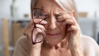 An older woman with her eyes closed in discomfort, rubbing them with her fingers while holding a pair of glasses in one hand.