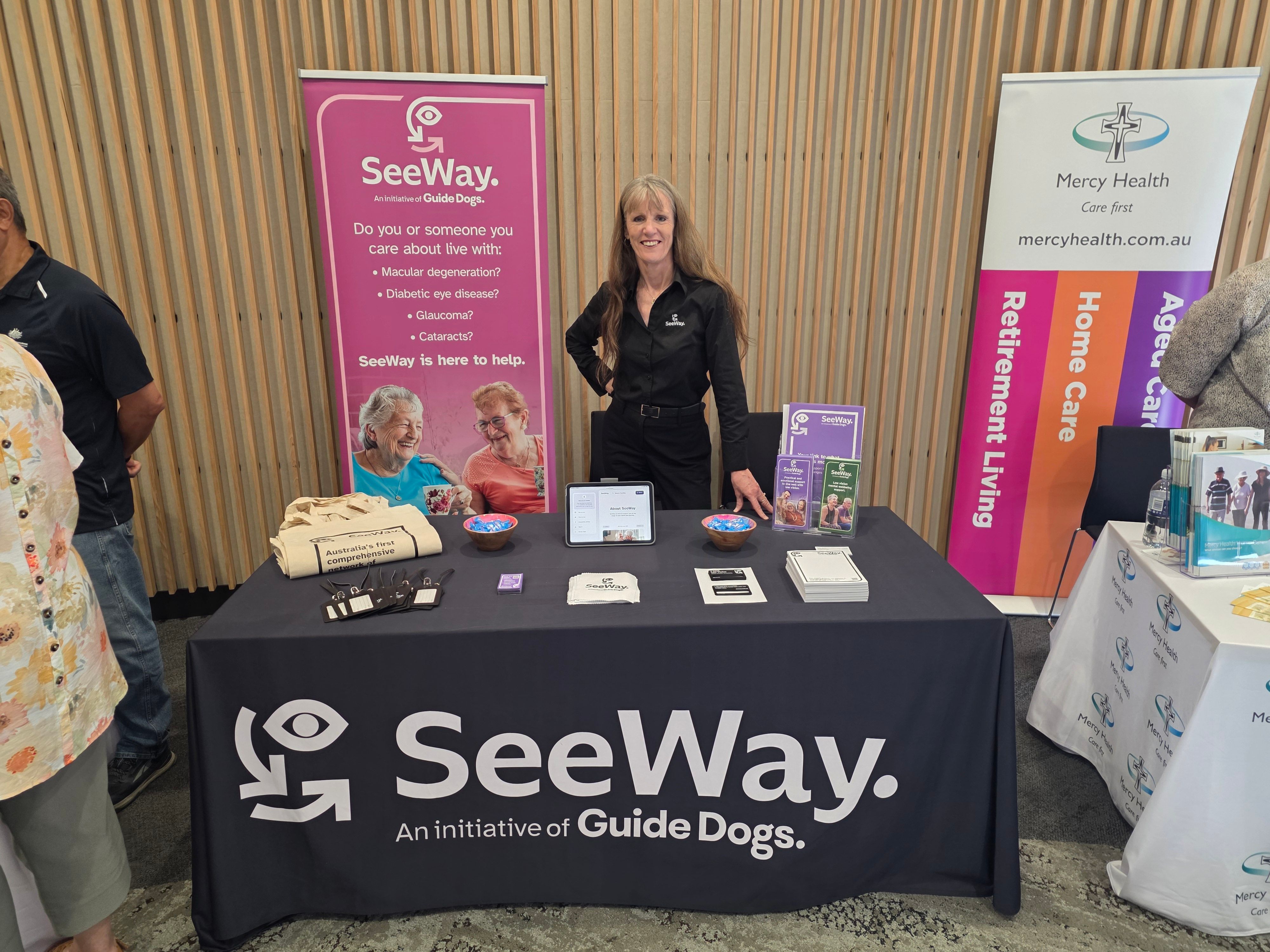 SeeWay team member Kaye standing behind a trade table at an expo. The table is covered in different SeeWay flyers and there is a SeeWay banner behind her.
