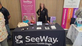 SeeWay team member Kaye standing behind a trade table at an expo. The table is covered in different SeeWay flyers and there is a SeeWay banner behind her.