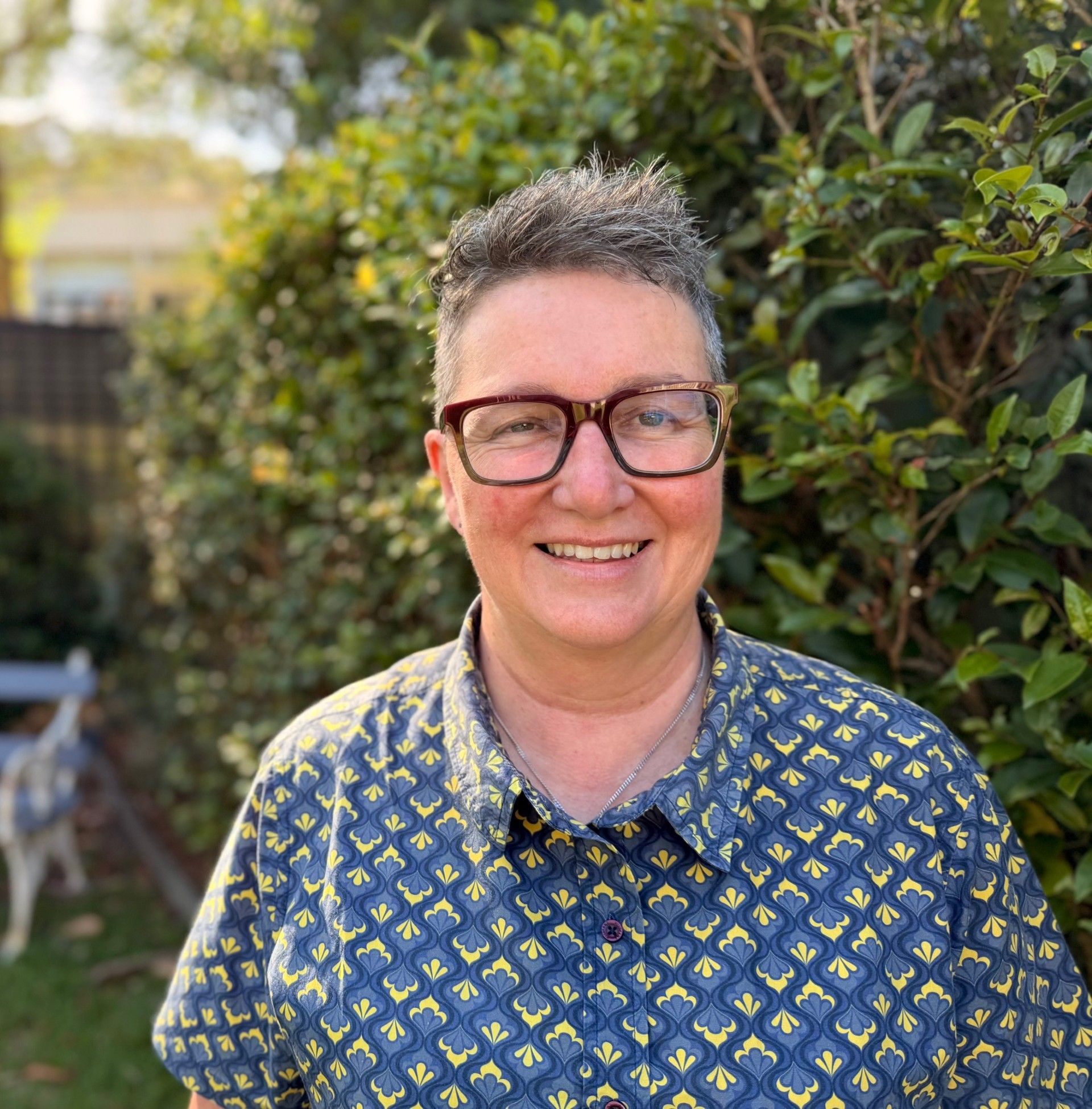 A photo of Nerida smiling at the camera standing in front of a hedge.