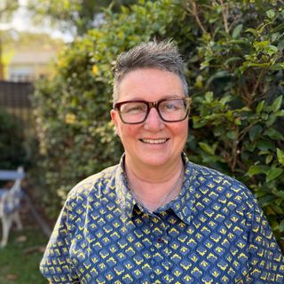 A photo of Nerida smiling at the camera standing in front of a hedge.