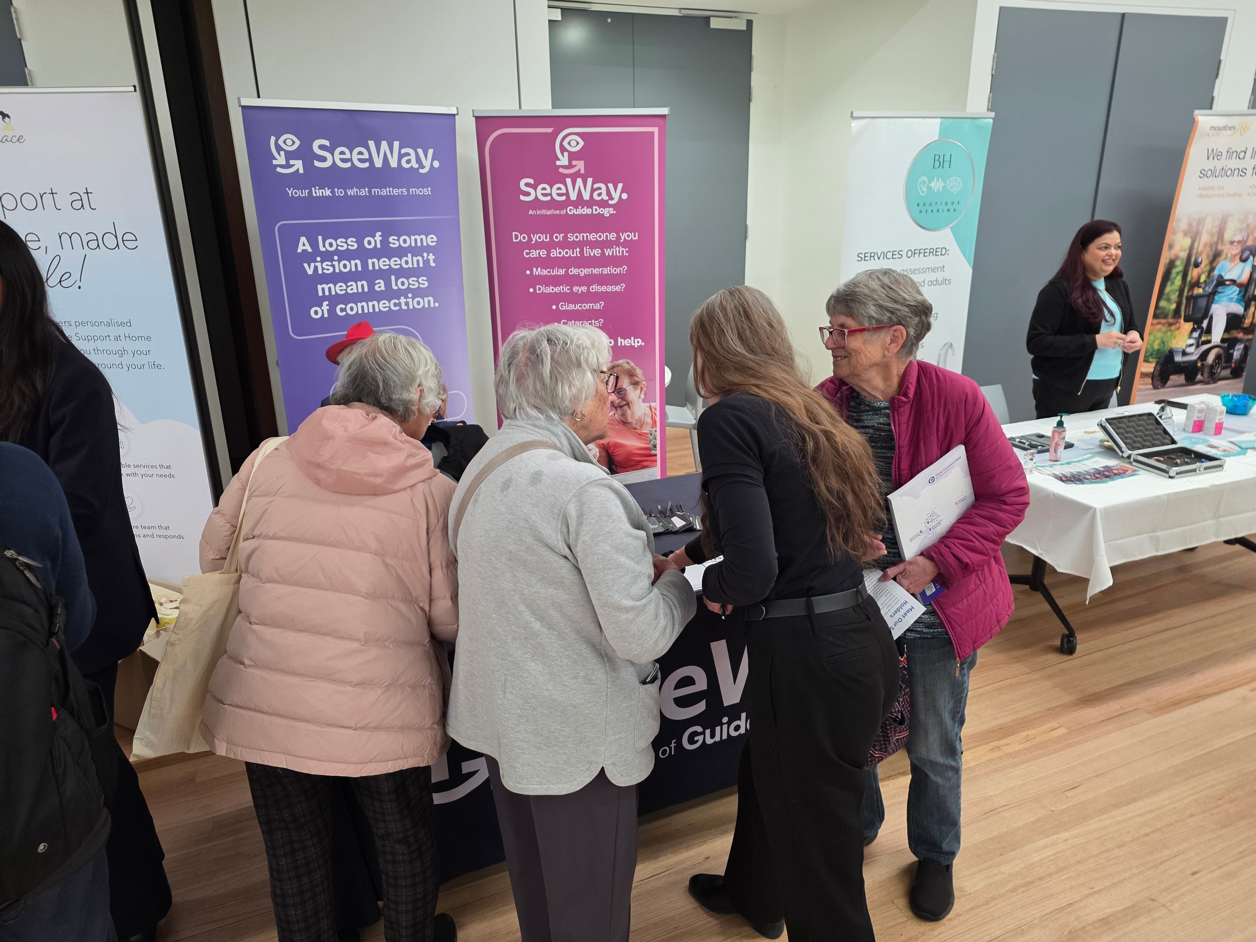 SeeWay team member Kaye talking with Clients at a community event, all with their backs to the camera as they look the SeeWay display table and banners.