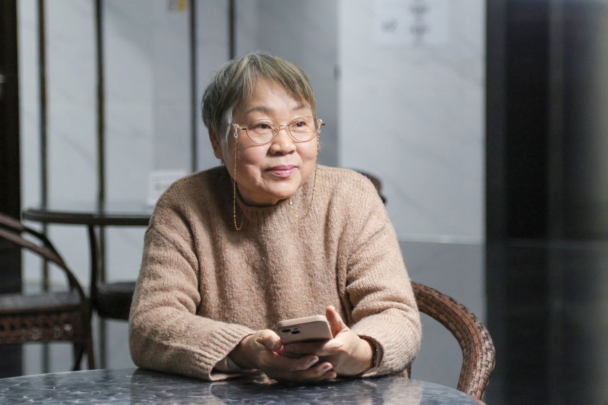 An older woman seated at a table holding a smartphone and looking into the distance.