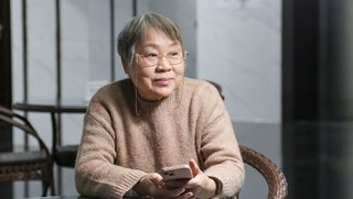 An older woman seated at a table holding a smartphone and looking into the distance.