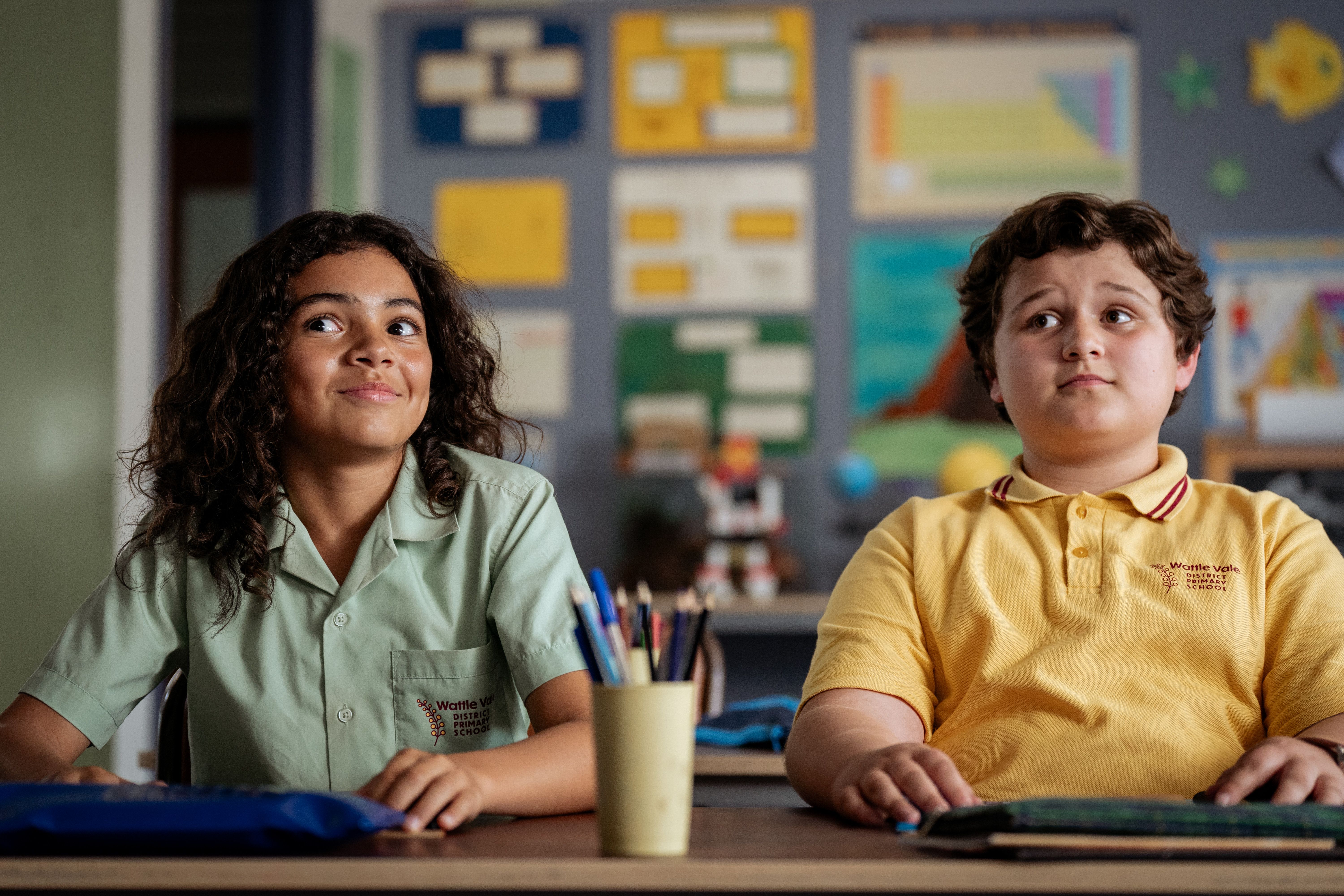 Two 10-year-old school students are sat at desks in a classroom, the cheeky boy on the left hides a barely contained smile.