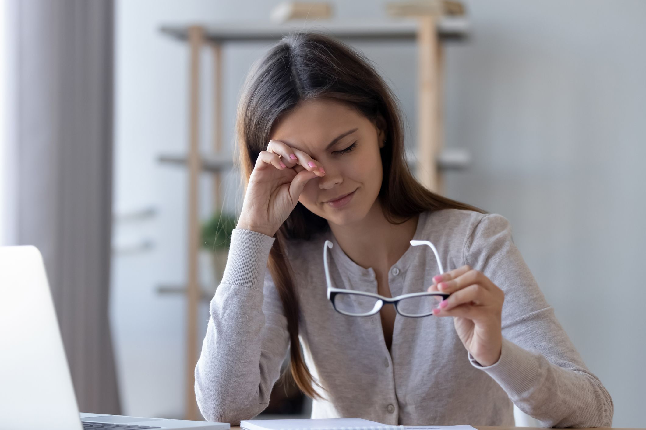 A woman sits at a computer desk, her spectacles are held in one hand, she rubs her eye with the other hand.