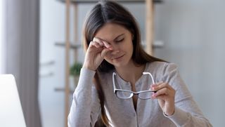 A woman sits at a computer desk, her spectacles are held in one hand, she rubs her eye with the other hand.