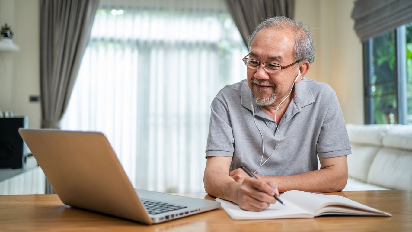 Older person using laptop with headphones while writing in a notebook