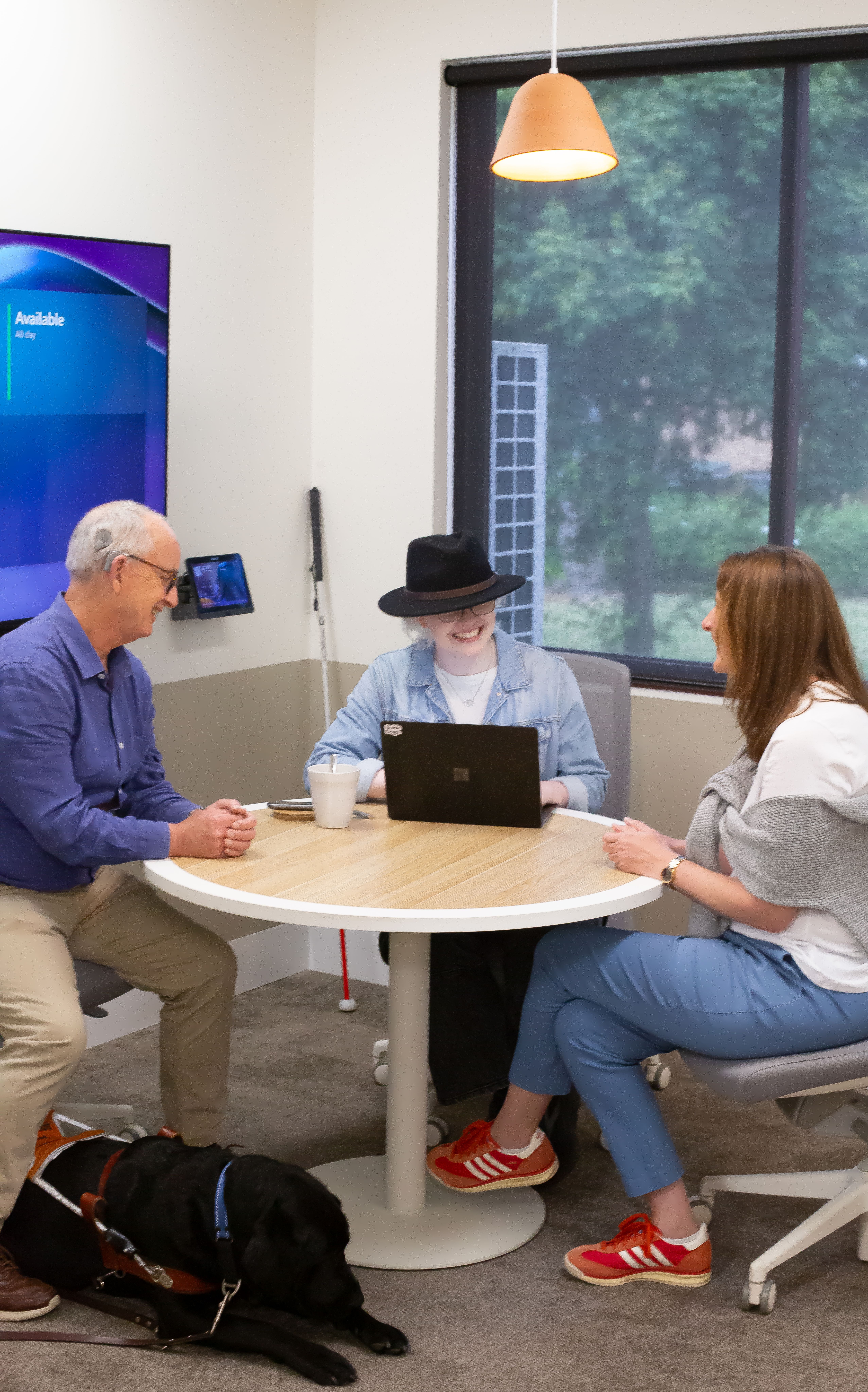Three colleagues meet at a table. A guide dog lies beside one and a white cane rests by another.