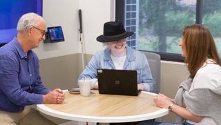 Three colleagues meet at a table. A guide dog lies beside one and a white cane rests by another.