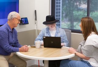 Three colleagues meet at a table. A guide dog lies beside one and a white cane rests by another.