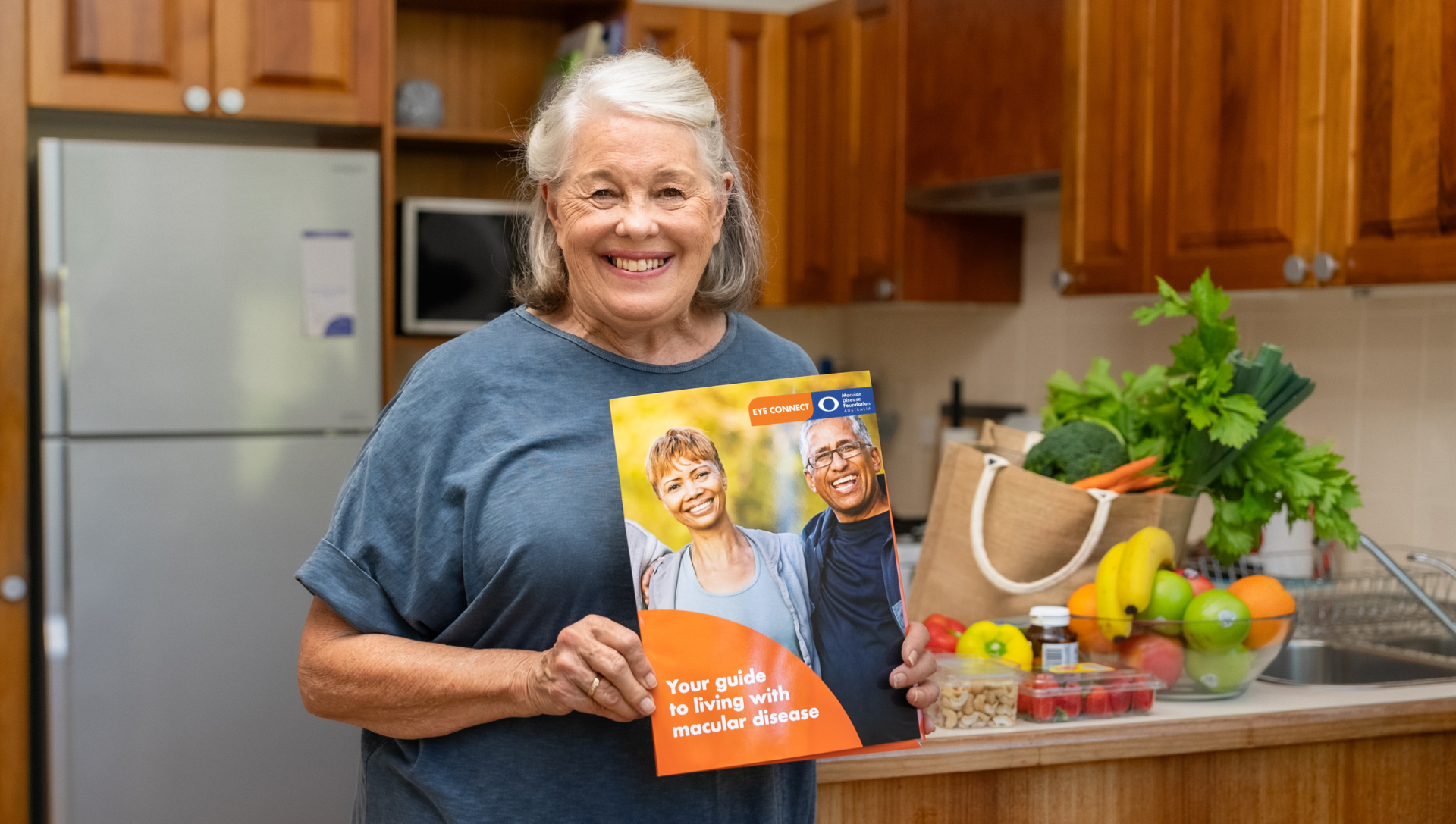 A smiling woman holding the Macular Disease Foundation newsletter in her kitchen.