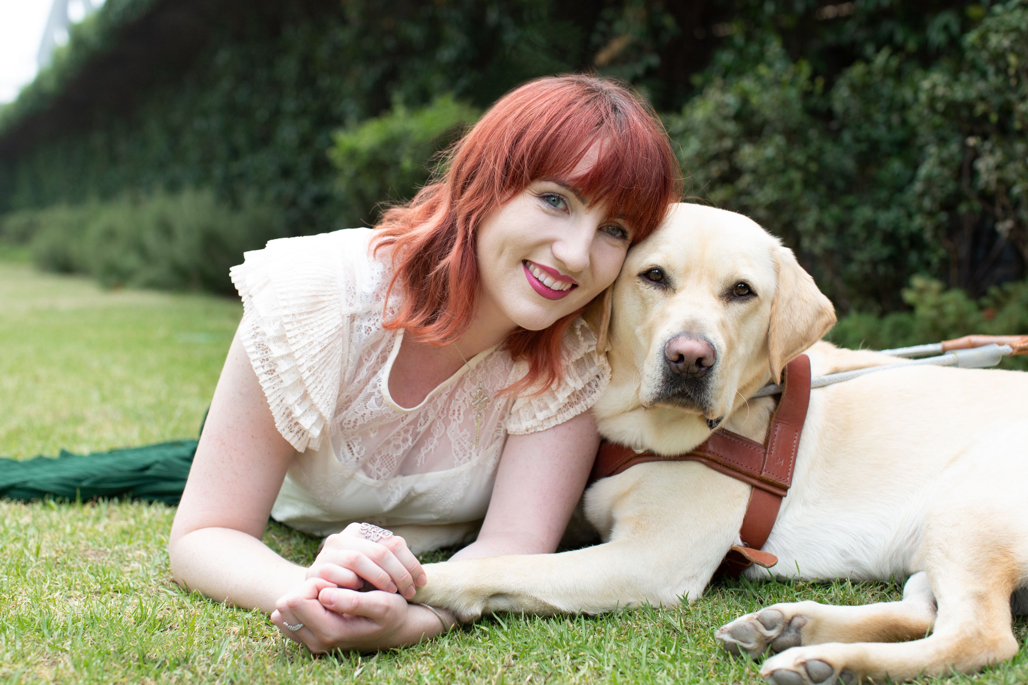 A smiling woman with red hair lying on the grass besides a yellow Labrador Guide Dog in a leather harness.