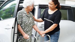 Carer helping older person using a walking frame to exit a car