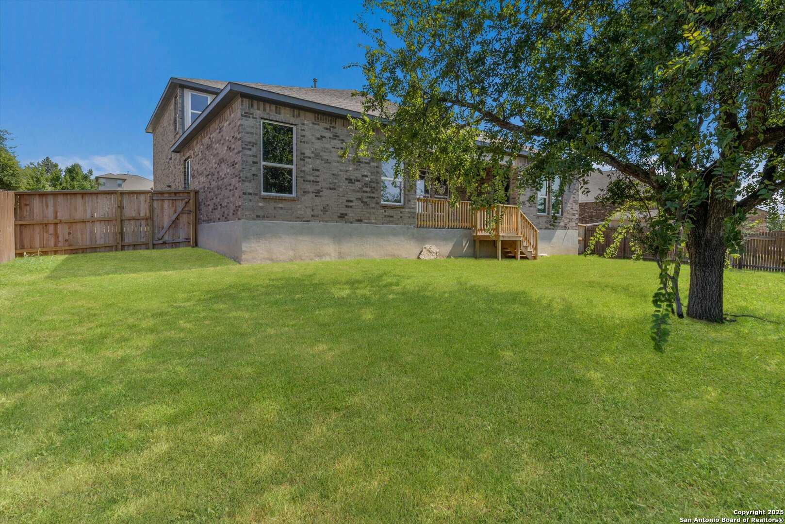 Two-story brick home backyard with wooden deck, mature oak tree, lush green lawn, and privacy fence in Ladera, San Antonio, Texas