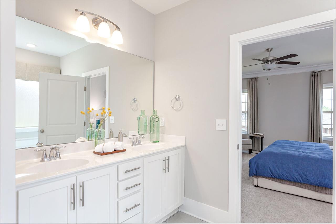 Spacious master bathroom in The Rockford with double vanity, large mirror, green bottles, and adjacent bedroom with blue bedding