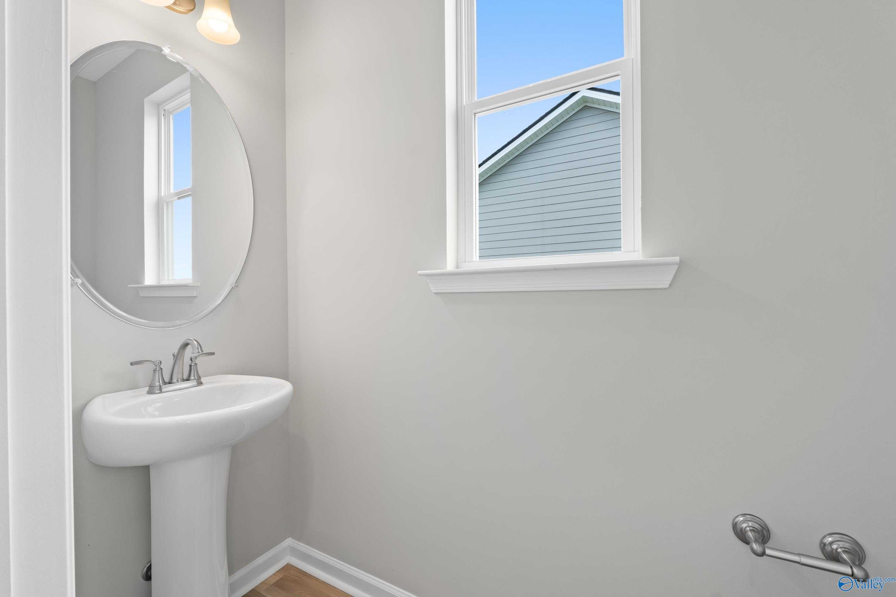 Elegant powder room with white pedestal sink, round mirror, and window in Evermore Homes The Augusta, Madison, Alabama