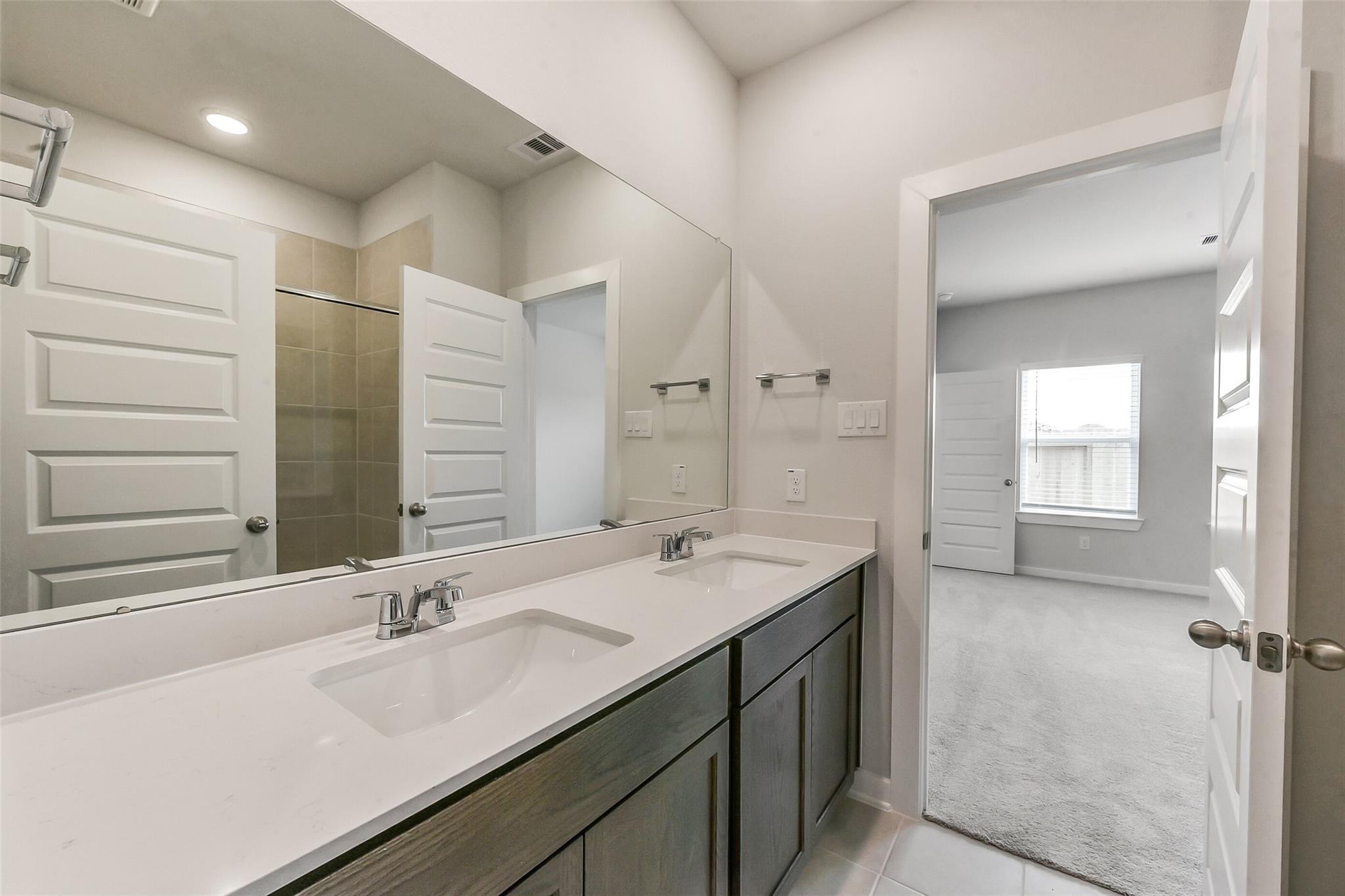 Modern master bathroom featuring double quartz vanity, gray cabinets, large mirror, and tiled shower in Davidson Homes The Sabine F, Dayton, Texas