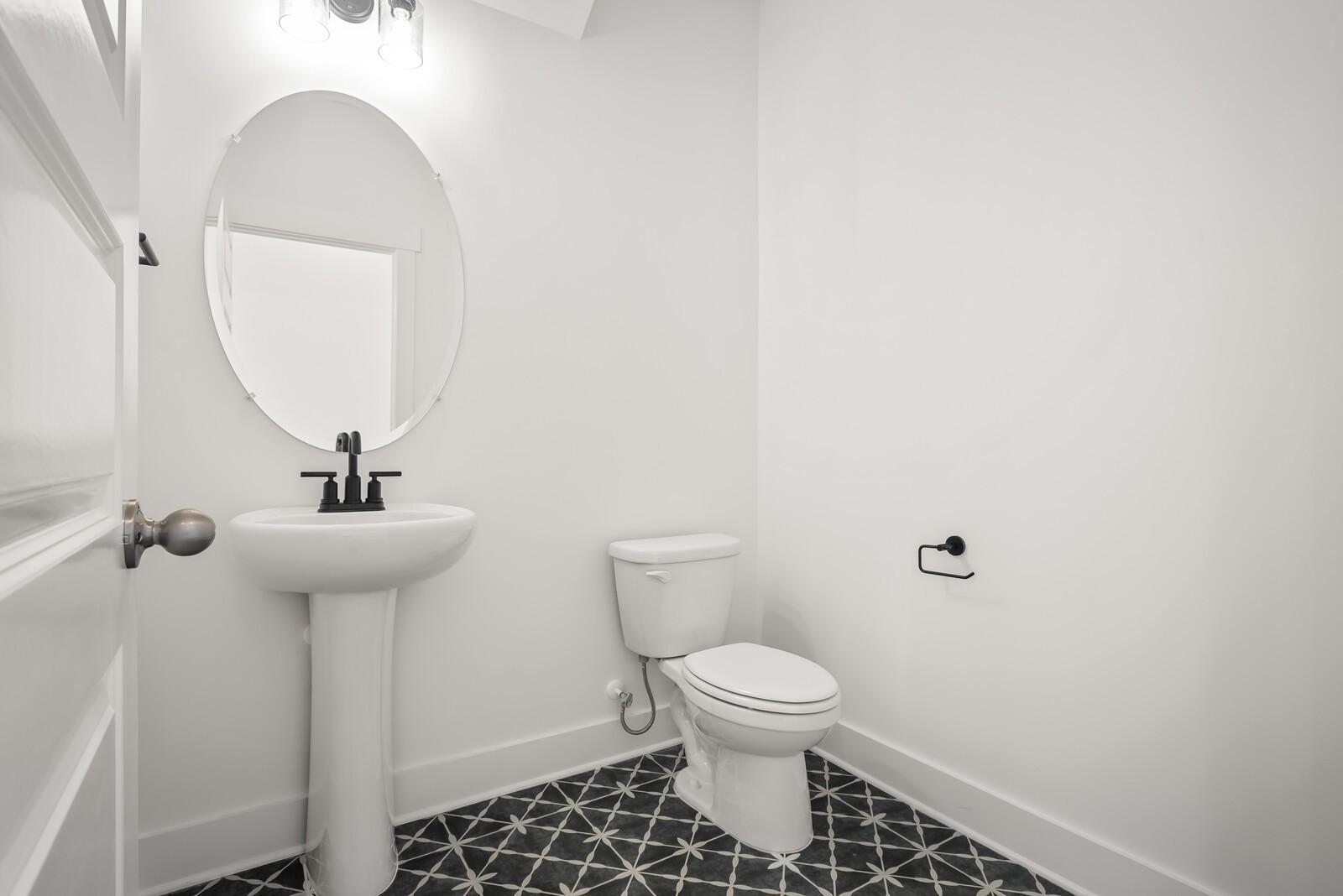 Modern powder room with white pedestal sink, oval mirror, toilet, and black geometric tile floor in Davidson Homes The Logan C, Gallatin, TN