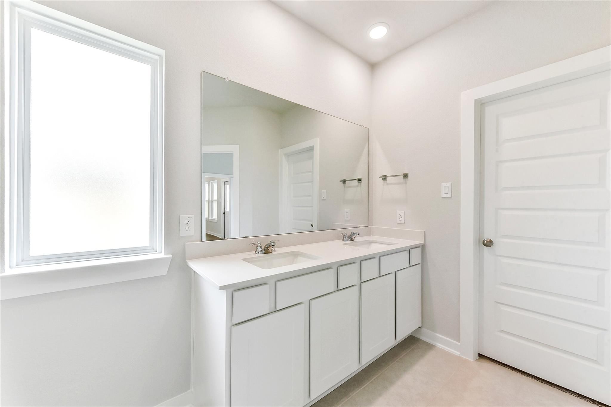 Modern master bathroom featuring double vanity with white cabinets, large mirror, and window in Davidson Homes The Costa B, Dayton, Texas
