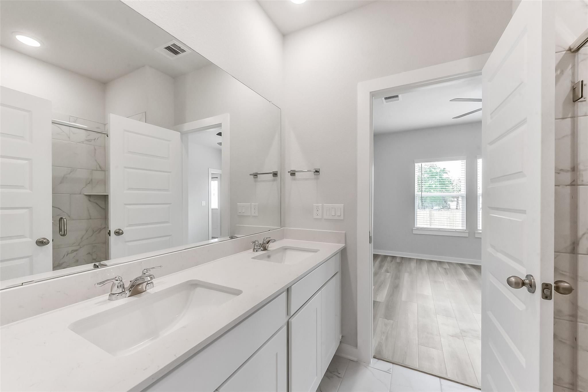 Modern master bathroom with double vanity, quartz counters, marble shower, and open bedroom door in Davidson Homes The Frio G, Magnolia Texas