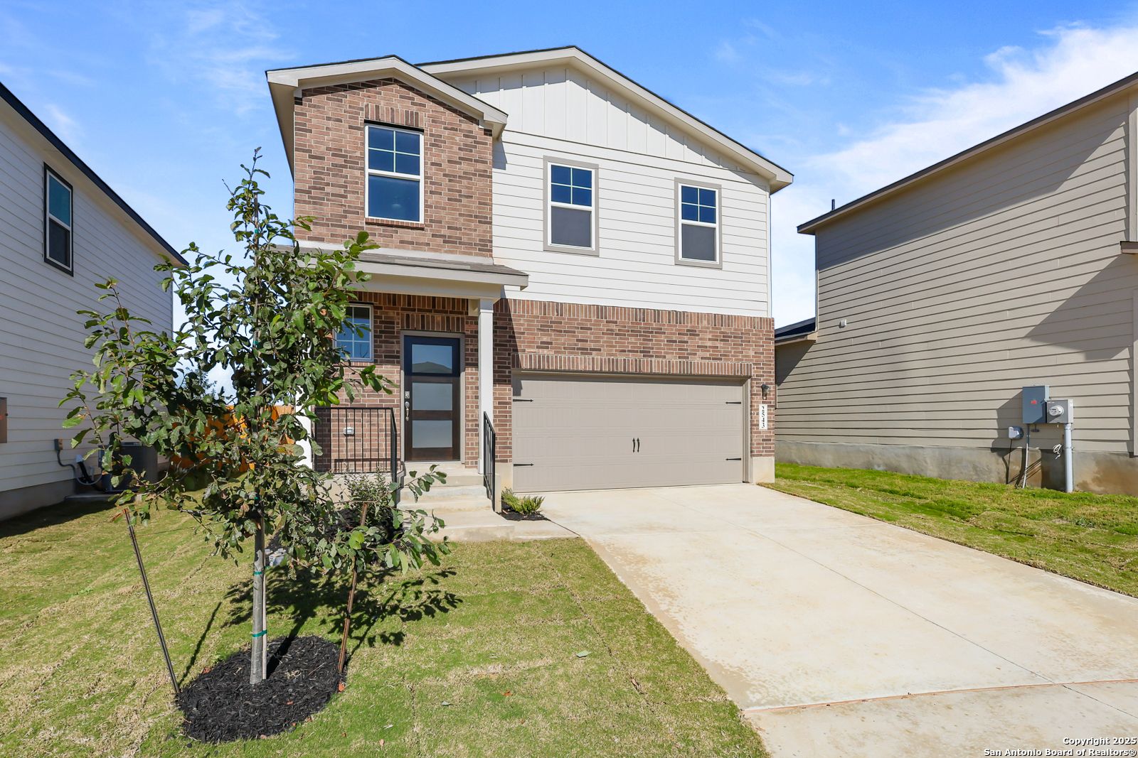 Modern two-story brick home with white siding, 2-car garage, front porch, and young tree in green yard, Applewhite Meadows, San Antonio