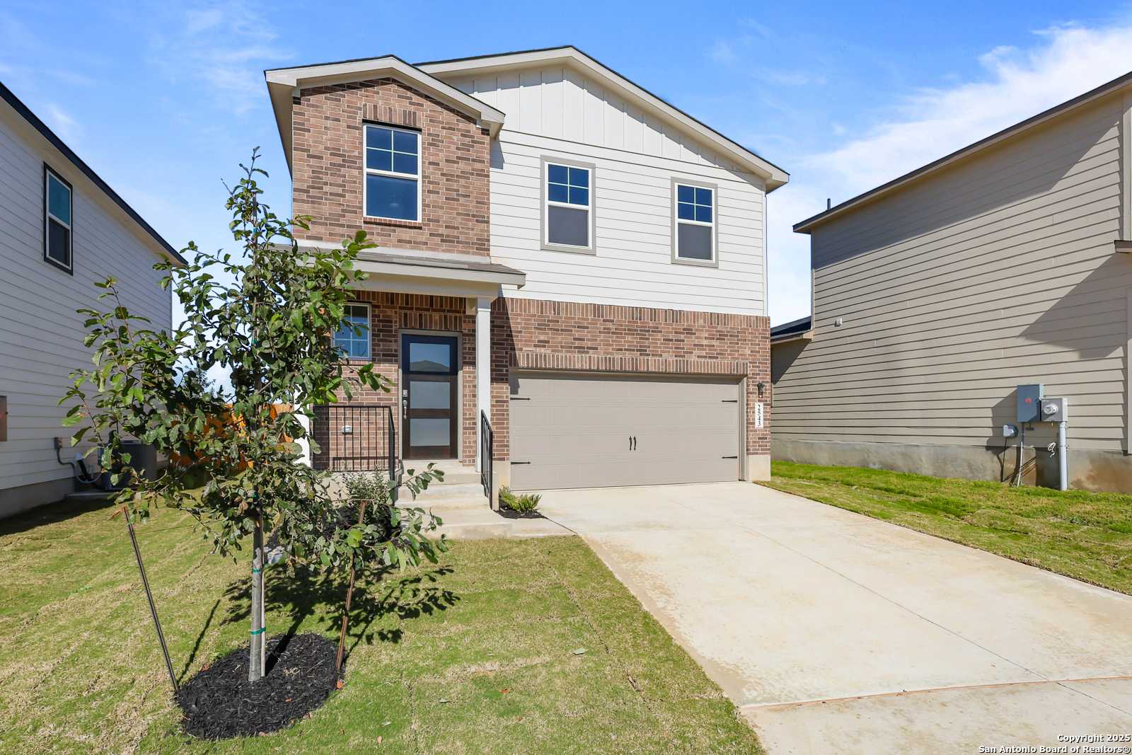 Modern two-story brick home with white siding, 2-car garage, front porch, and young tree in green yard, Applewhite Meadows, San Antonio