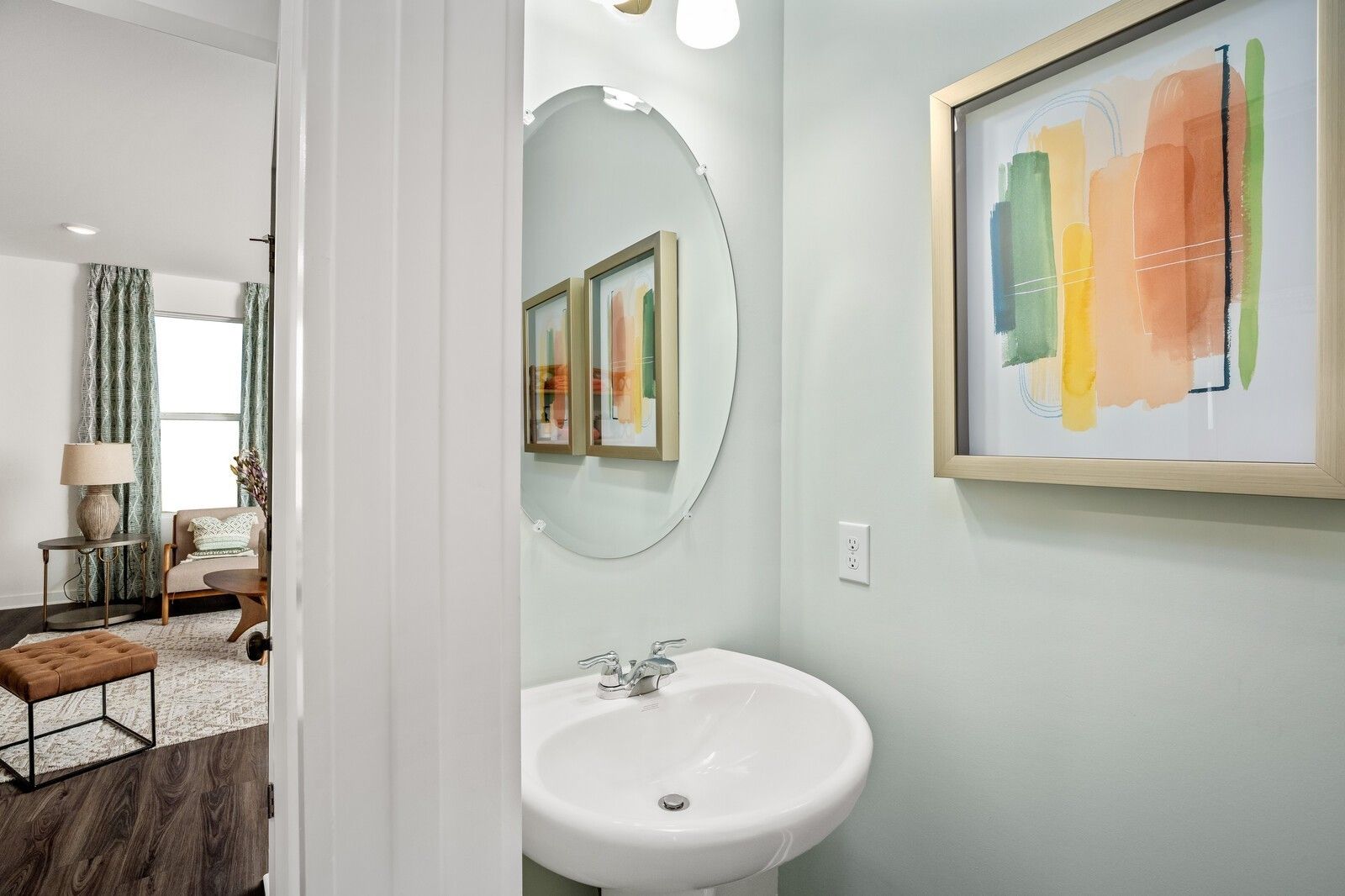 Elegant powder room with white pedestal sink, round mirror, abstract art on soft green walls, open to living area in Davidson Homes The Gordon C, White House, TN