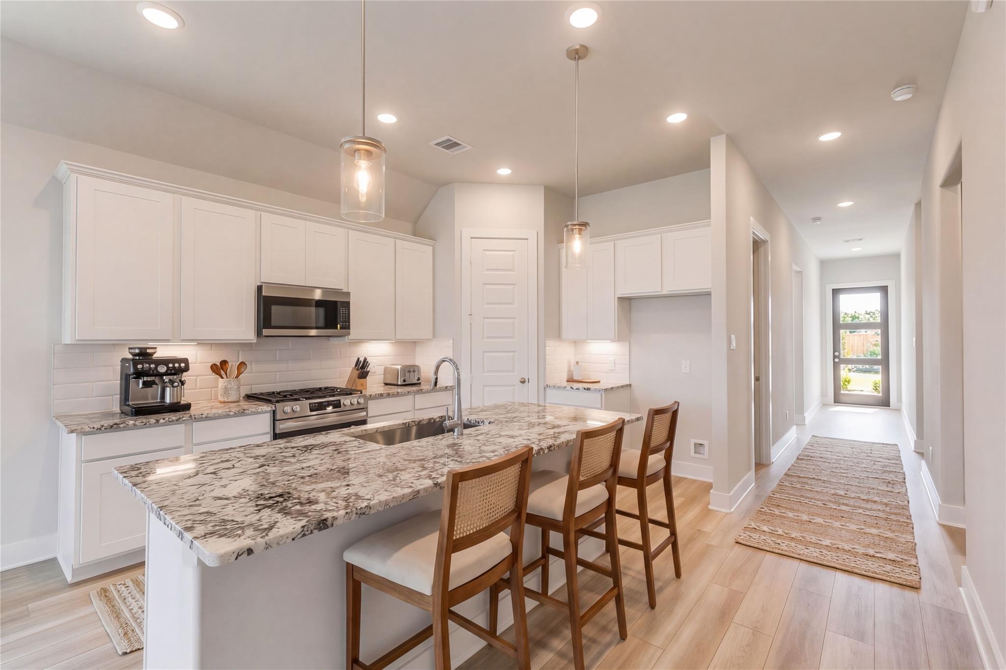 Modern kitchen with white cabinets, speckled granite island, stainless steel appliances, and rattan bar stools in The Luna L, Sunterra, Katy, Texas