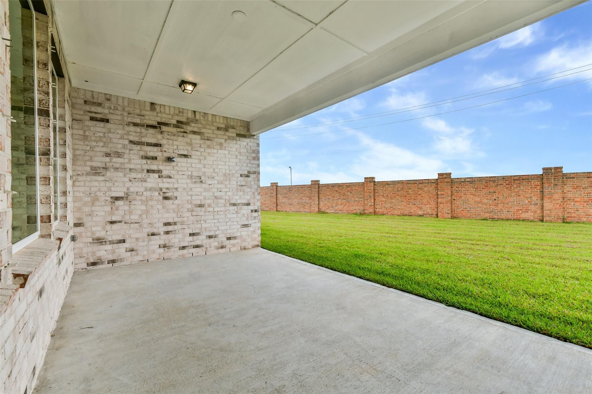 Covered back patio with white brick walls, concrete flooring, lush green lawn, and red brick fence in Lago Mar, Texas City home