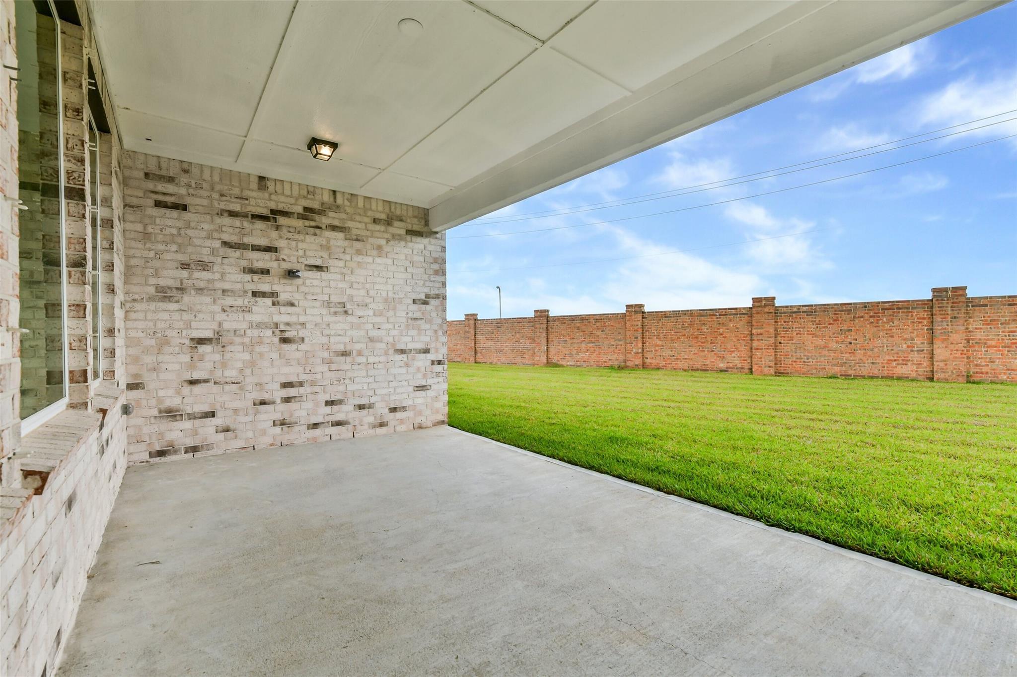 Covered back patio with white brick walls, concrete flooring, lush green lawn, and red brick fence in Lago Mar, Texas City home