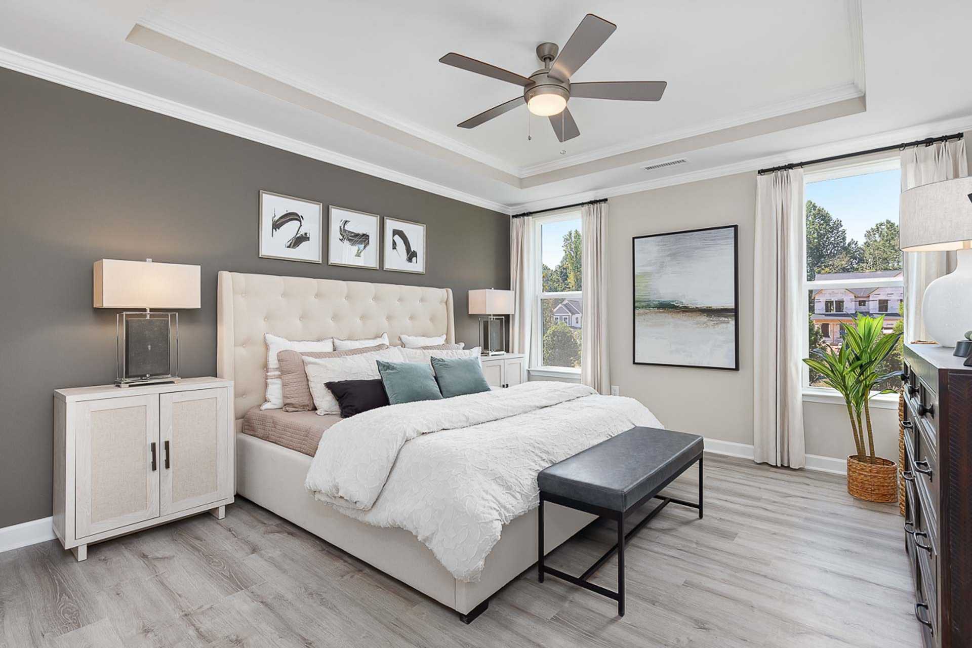 Spacious primary bedroom in Prince Place Fuquay-Varina NC with king bed, tufted headboard, ceiling fan, wall art, and hardwood floors