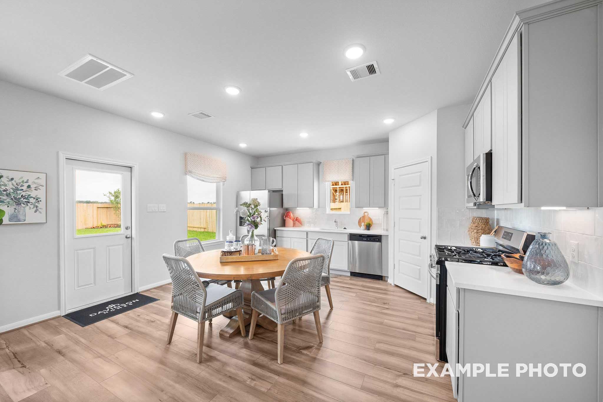 Open-concept dining area in The Trinity E with round wood table, white shaker cabinets, stainless appliances, and backyard view