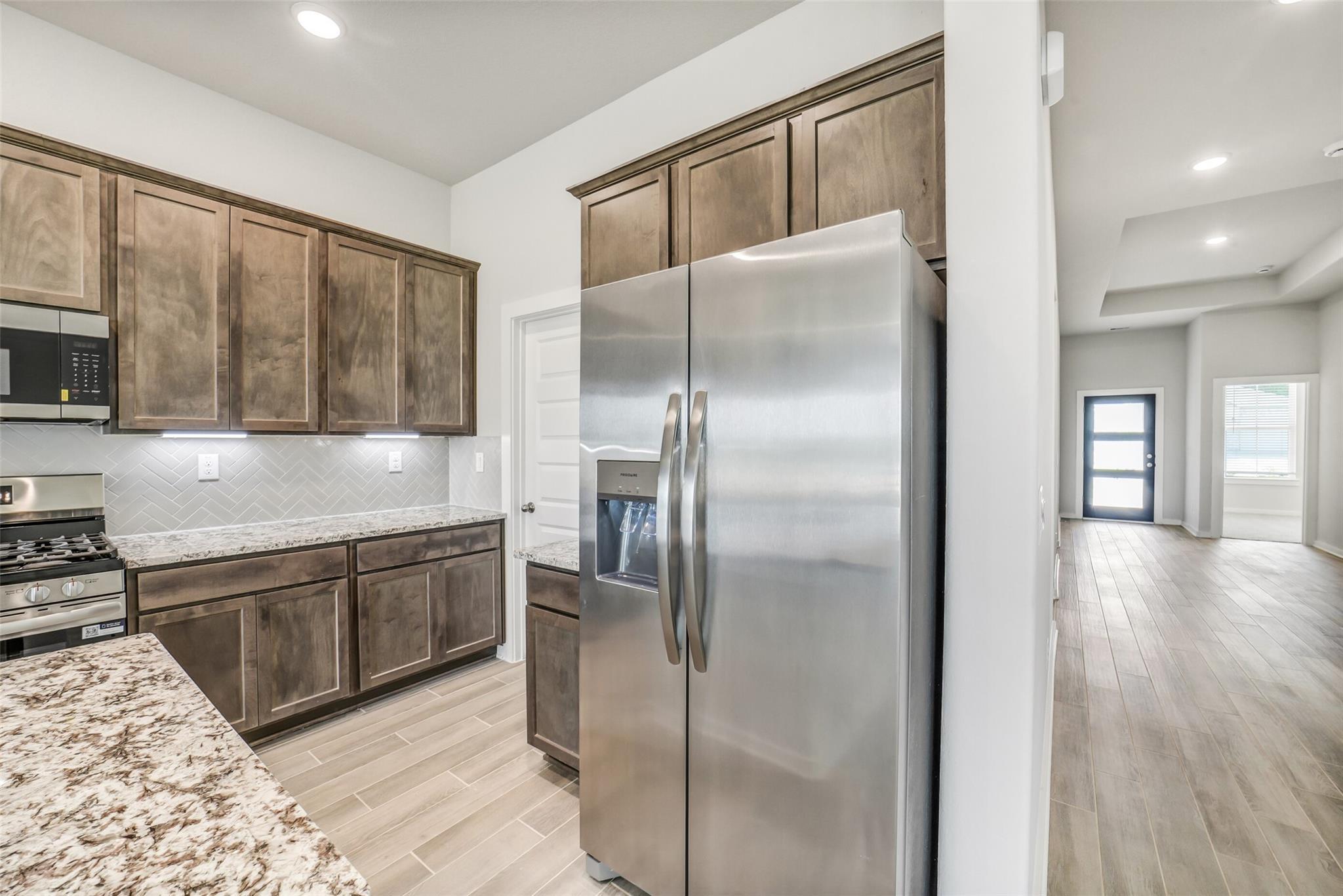 Modern kitchen featuring shaker cabinets, granite countertops, stainless steel fridge, and herringbone backsplash in The Acadia C, Magnolia, TX