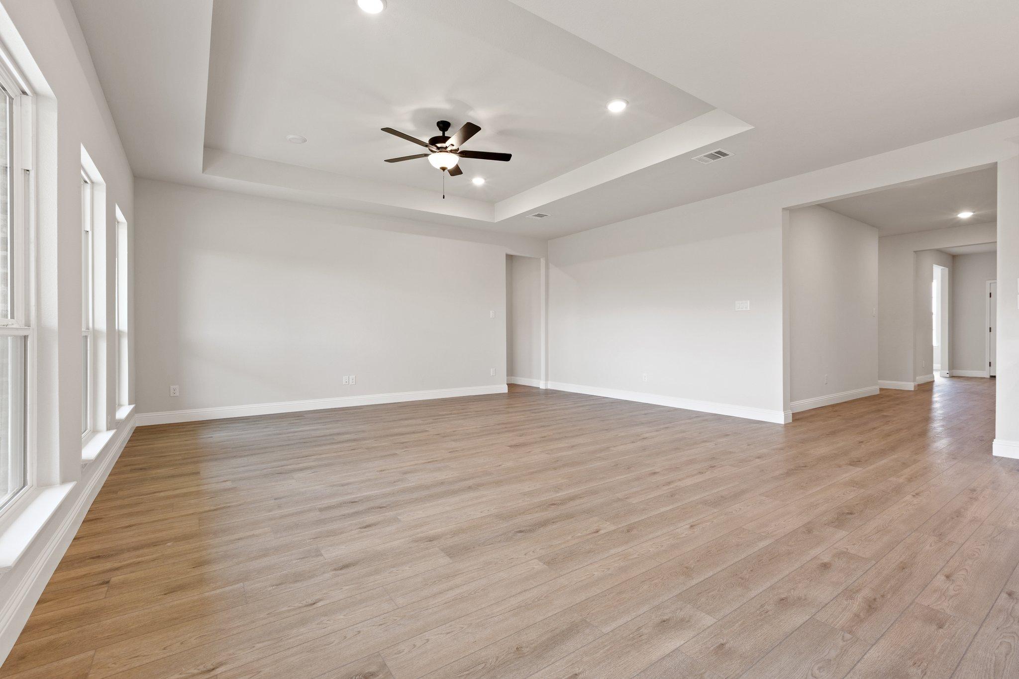 Spacious open living area in The Harrison with light hardwood floors, tray ceiling fan, white walls, and large windows