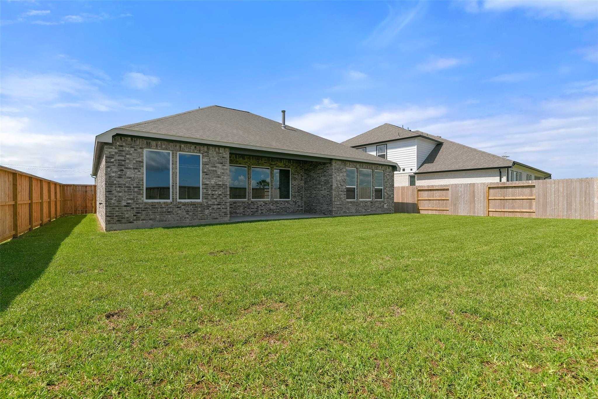 Single-story brick home backyard with large windows, covered patio, lush green lawn, and wooden fence in Lago Mar, Texas City