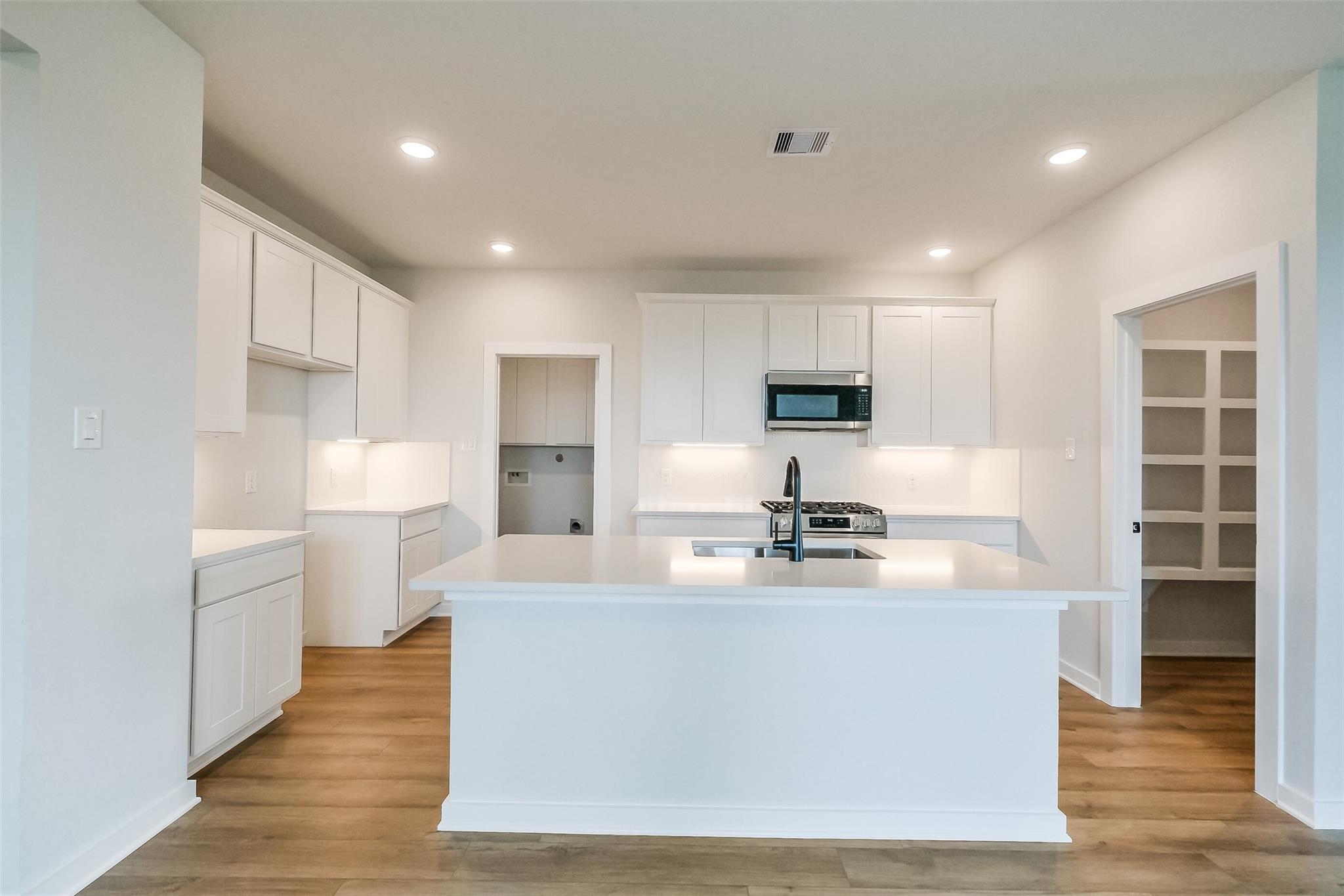 Modern white kitchen with island sink, stainless appliances, and hardwood floors in Davidson Homes The Tierra B, Beasley, Texas